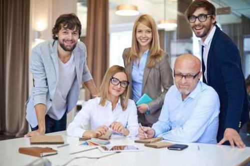Das Team von eXpletus - Fünfköpfiges Team in Businesskleidung sitzt und steht gemeinsam an einem Tisch in einem modernen Büro, mit Laptops, Notizbüchern und Unterlagen vor sich